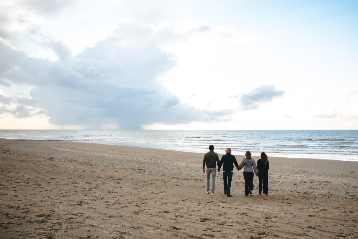Gezin van vier wandelt hand in hand op het strand, gezien van achter met bewolkte lucht — familiefotografie door Nanou Verhoeven