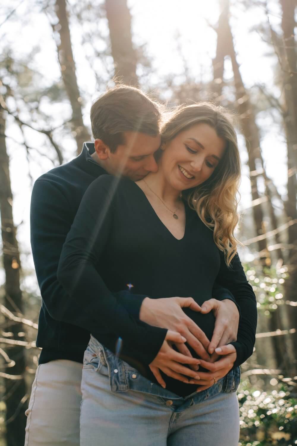 Zwangere vrouw en partner omhelzen elkaar in het bos, handen op de buik — zwangerschapsfotografie door Nanou Verhoeve
